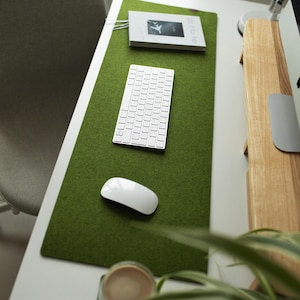 May include: A green felt desk mat with a white keyboard, a white mouse, and a book with the text "WORK" on the cover. The mat is on a white desk with a wooden monitor stand.
