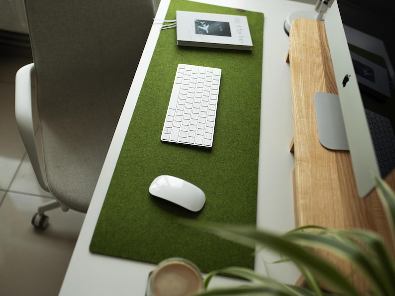 May include: A green felt desk mat with a white keyboard, a white mouse, and a book with the text "WORK" on the cover. The mat is on a white desk with a wooden monitor stand.