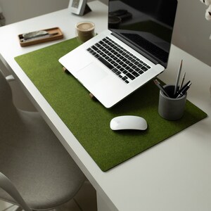May include: A green felt desk mat with a white wireless mouse, a laptop computer, and a gray pencil holder with pens on a white desk.