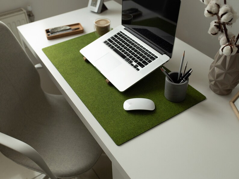 May include: A green felt desk mat with a white wireless mouse, a laptop computer, and a gray pencil holder with pens on a white desk.