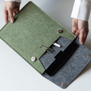 May include: A sage green and charcoal gray felt document sleeve featuring a fold-over flap with two silver-toned magnetic snap closures. The interior contains a black tablet, a white stylus, and two metallic pen-like tools held in place.