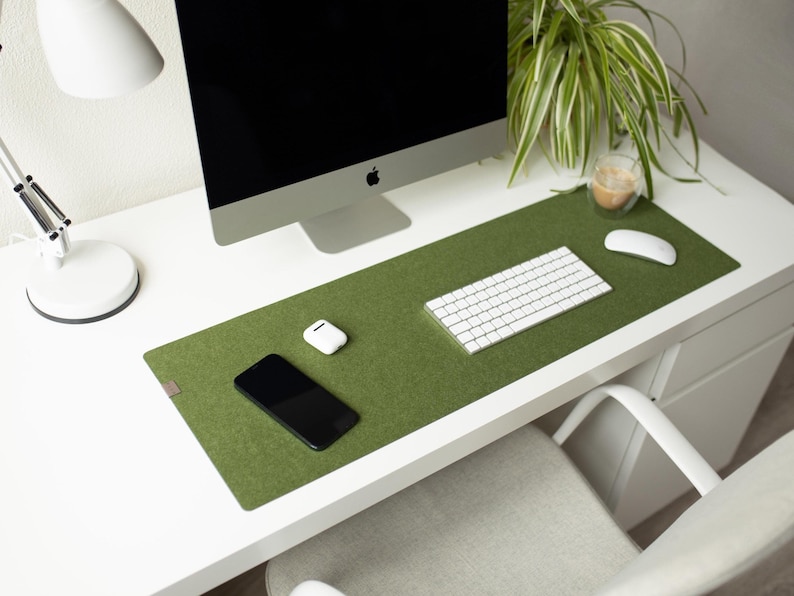May include: A green felt desk mat with a white keyboard, a white mouse, a white phone, and a white wireless charging pad. The mat is on a white desk with a white lamp and a plant in the background.