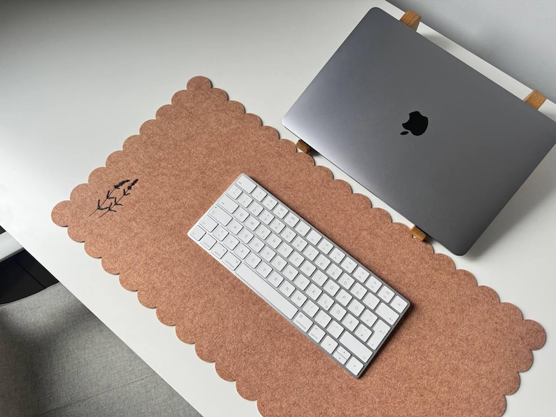 Puede incluir: Un espacio de trabajo inform&aacute;tico con un port&aacute;til gris, un teclado blanco y una alfombrilla de escritorio de fieltro marr&oacute;n con borde festoneado y dise&ntilde;o de ramita de lavanda. El conjunto est&aacute; sobre un escritorio blanco.