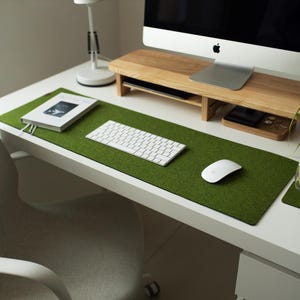 May include: A green felt desk mat with a white keyboard, mouse, and a book on top. The mat is on a white desk with a wooden monitor stand and a cup of coffee on a coaster.