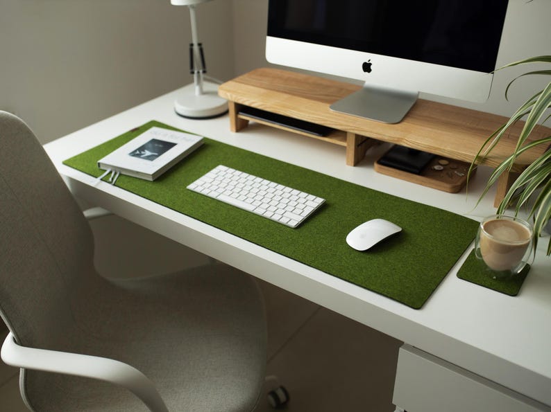 May include: A green felt desk mat with a white keyboard, mouse, and a book on top. The mat is on a white desk with a wooden monitor stand and a cup of coffee on a coaster.