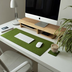 May include: A white computer monitor on a wooden stand with a white keyboard and mouse on a green felt desk mat. A cup of coffee sits on a green felt coaster on a white desk.