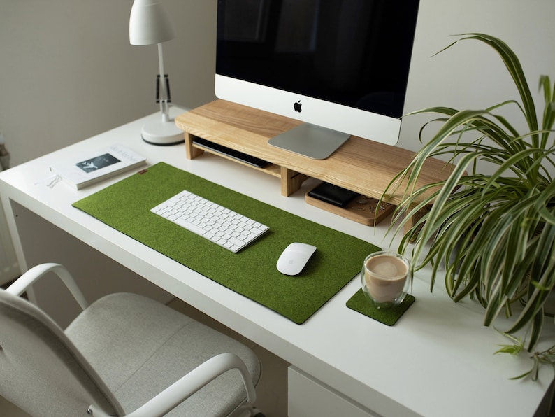 May include: A white computer monitor on a wooden stand with a white keyboard and mouse on a green felt desk mat. A cup of coffee sits on a green felt coaster on a white desk.