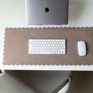 以下が含まれることがあります： 上から見たワークスペース。ラップトップ、キーボード、マウスが、スカラップエッジの明るい茶色のフェルトデスクマットの上に配置されています。デスクは白で、灰色の椅子が見えます。