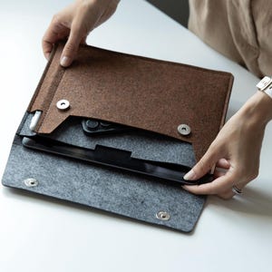 May include: A brown and gray felt laptop sleeve with a black tablet and a phone inside. The sleeve has a flap closure with silver snap buttons. The person is holding the sleeve open.