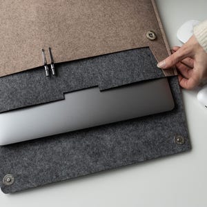 Könnte beinhalten: Eine zweifarbige Laptop-Hülle aus Filz mit einer beigen Außenklappe und einem dunkelgrauen Innenfach. Die Hülle verfügt über zwei silberne Metalldruckknöpfe als Verschluss sowie einen Schlitz für einen Stift und enthält einen silbernen Laptop.