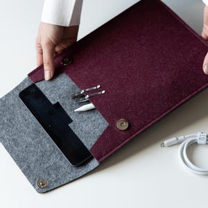 May include: A burgundy and gray felt sleeve for a tablet, featuring a fold-over flap with two silver-toned magnetic snap closures. The interior contains a black tablet, a white stylus, and two silver metal clips. A white charging cable is nearby.