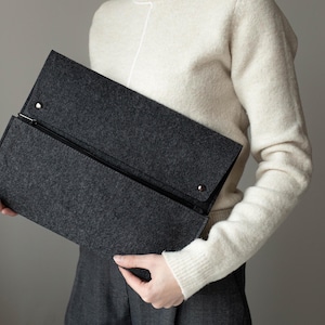 May include: A gray felt clutch with a zipper closure. The clutch is held by a person wearing a white sweater.