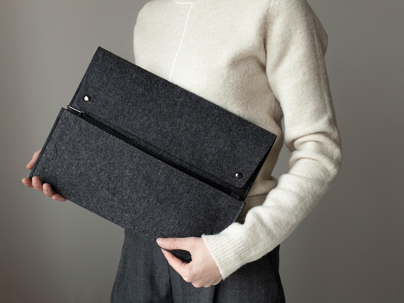 May include: A gray felt clutch with a zipper closure. The clutch is held by a person wearing a white sweater.