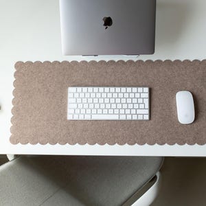 Puede incluir: Un espacio de trabajo inform&aacute;tico con un port&aacute;til gris, un teclado y un rat&oacute;n blancos sobre una alfombrilla de escritorio marr&oacute;n con borde festoneado. Un libro es visible a la izquierda. La configuraci&oacute;n est&aacute; sobre un escritorio blanco.