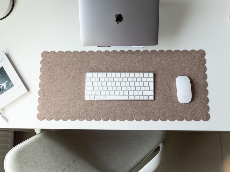 Puede incluir: Un espacio de trabajo inform&aacute;tico con un port&aacute;til gris, un teclado y un rat&oacute;n blancos sobre una alfombrilla de escritorio marr&oacute;n con borde festoneado. Un libro es visible a la izquierda. La configuraci&oacute;n est&aacute; sobre un escritorio blanco.