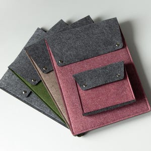 Può includere: Una pila di quattro custodie rettangolari per documenti in feltro con patte grigio scuro fissate da bottoni a pressione in metallo argentato. Le custodie presentano diversi colori di base tra cui grigio scuro, verde oliva, beige e magenta.