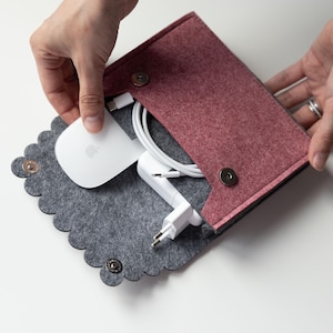 May include: A felt storage pouch with a scalloped edge and a maroon flap featuring two metallic snap buttons. The pouch contains a white computer mouse, a white power adapter, and a white charging cable, all organized within the gray felt interior.