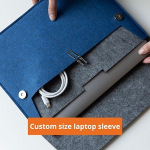 Könnte beinhalten: Eine Laptoptasche aus blauem und grauem Filz mit einem Klappverschluss und zwei silberfarbenen Magnetknöpfen. Im Inneren befinden sich ein weißes Ladekabel und ein dunkelgrauer Laptop. Der Text "Custom size laptop sleeve" steht auf einem orangefarbenen Balken.