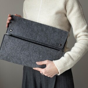 May include: A gray felt clutch with a zipper closure and a flap with a snap closure. The clutch is held by a person wearing a white sweater.