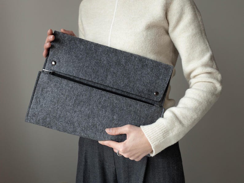 May include: A gray felt clutch with a zipper closure and a flap with a snap closure. The clutch is held by a person wearing a white sweater.