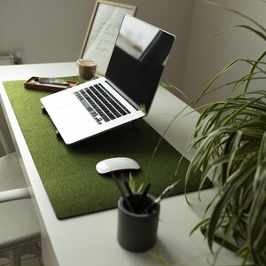 May include: A white laptop computer with a green felt desk mat on a white desk. The laptop is open and the screen is black. There is a plant in the background.