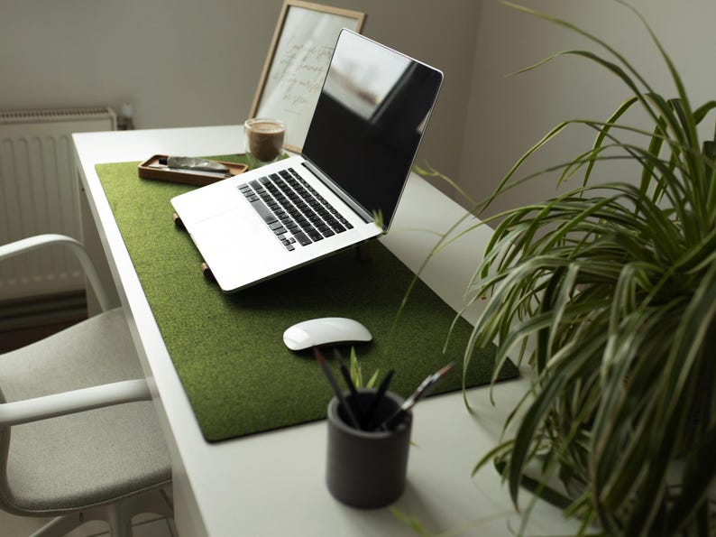 May include: A white laptop computer with a green felt desk mat on a white desk. The laptop is open and the screen is black. There is a plant in the background.
