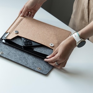 May include: A felt laptop sleeve in tan and gray, with a black tablet and smartphone inside. The sleeve is open, revealing the contents. The design includes snap closures and a cutout for the devices.