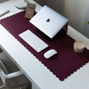 Puede incluir: Una alfombrilla de escritorio rectangular de fieltro color burdeos oscuro con un borde festoneado. La alfombrilla se coloca sobre una superficie blanca y sirve de base para un portátil plateado sobre un soporte, un teclado blanco y un ratón.
