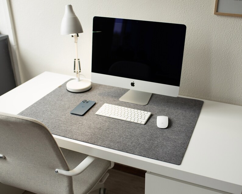 May include: A white desk with a gray felt desk mat, a white lamp, a computer monitor, a keyboard, a mouse, and a smartphone.