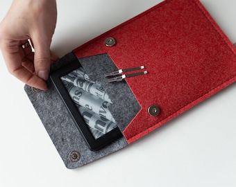 May include: A red and grey felt sleeve case with magnetic snap closures, containing a black e-reader device with a screen displaying newspaper-style text. Two small silver metal stylus tools are resting on the grey felt interior flap.