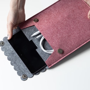 Op de afbeelding: Een tweekleurige vilten hoes met een oudroze buitenkant en een grijze binnenkant met een geschulpte kleprand. De hoes bevat een zwarte tablet, een witte stylus en een witte oplaadkabel, vastgezet met twee ronde metalen drukknopen.