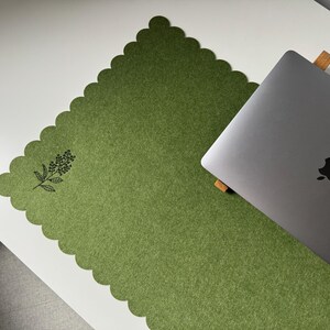 Puede incluir: Una alfombrilla de escritorio de fieltro verde con borde festoneado y dise&ntilde;o floral negro. Un port&aacute;til plateado descansa sobre la alfombrilla, colocada sobre un escritorio blanco. La alfombrilla proporciona una superficie suave.