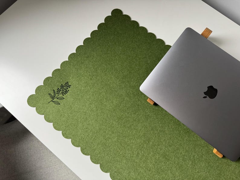 Puede incluir: Una alfombrilla de escritorio de fieltro verde con borde festoneado y dise&ntilde;o floral negro. Un port&aacute;til plateado descansa sobre la alfombrilla, colocada sobre un escritorio blanco. La alfombrilla proporciona una superficie suave.