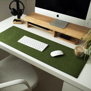 May include: A white desk with a green felt desk mat, a white keyboard, a white mouse, and a cup of coffee. The desk also has a wooden monitor stand with a computer monitor on top.