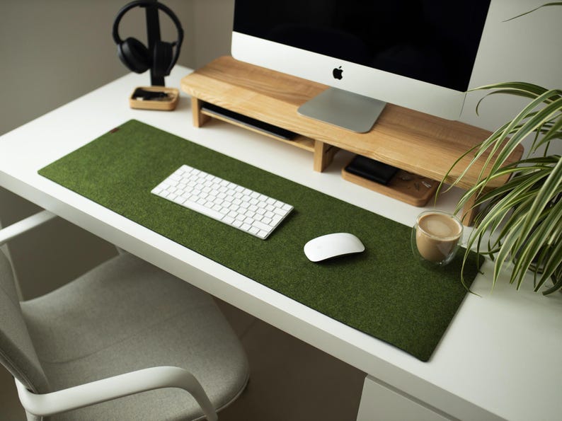 May include: A white desk with a green felt desk mat, a white keyboard, a white mouse, and a cup of coffee. The desk also has a wooden monitor stand with a computer monitor on top.