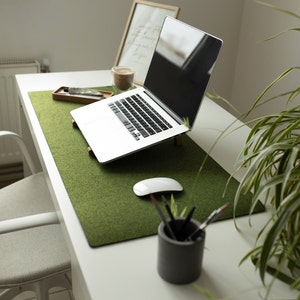 May include: A white laptop computer with a green felt desk mat on a white desk. A white mouse and a cup of pencils are on the desk. A houseplant is in the background.