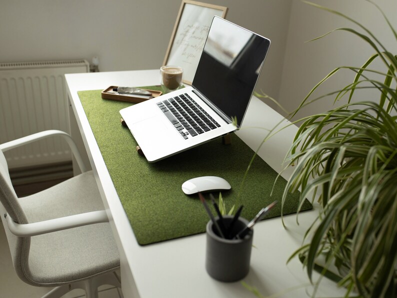 May include: A white laptop computer with a green felt desk mat on a white desk. A white mouse and a cup of pencils are on the desk. A houseplant is in the background.