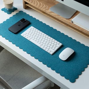 May include: A rectangular teal felt desk mat with a scalloped edge design. The mat is positioned on a white surface and holds a black smartphone, a white wireless keyboard, and a white computer mouse. A matching small scalloped felt coaster is included.