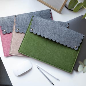 Könnte beinhalten: Drei rechteckige Laptop-Hüllen aus Filz in Grün, Beige und Rosa, jeweils mit einer dunkelgrauen, wellenförmig ausgeschnittenen Klappe, die mit einem silbernen Metalldruckknopf verschlossen wird. Die Hüllen sind gestapelt angeordnet.
