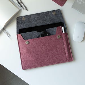 May include: A burgundy felt tablet sleeve with a gray interior, open to reveal a tablet, phone, and stylus. The sleeve has a snap closure and is next to a white mouse and a notebook. A pen and a card are also visible.
