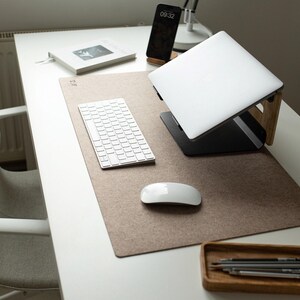 May include: A rectangular tan felt desk mat placed on a white desk. The mat holds a white wireless keyboard, a white wireless mouse, a silver laptop on a black and wood stand, a smartphone on a small wood stand, a book, and a wood tray with pens.