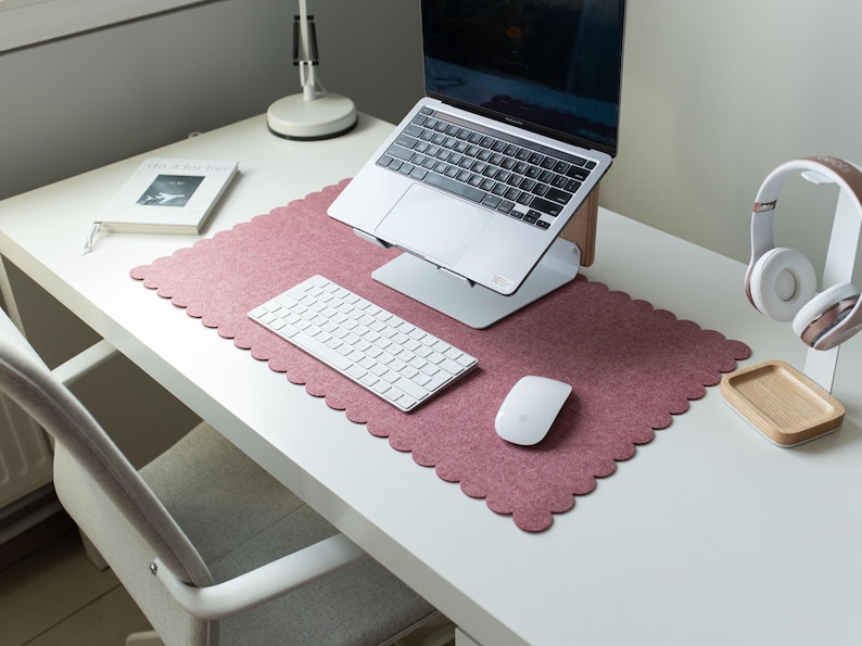 Puede incluir: Una configuraci&oacute;n de escritorio moderna con un port&aacute;til sobre un soporte, un teclado blanco y un rat&oacute;n. Una alfombrilla de escritorio de fieltro rosa con borde festoneado se encuentra debajo del ordenador y los accesorios. Tambi&eacute;n hay un cuaderno y auriculares.