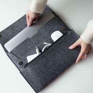 May include: A dark gray felt laptop sleeve with a fold-over flap secured by two dark metal snap buttons. The interior features a large compartment holding a silver laptop and a separate zippered pocket containing a white computer mouse and cables.