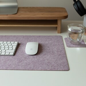 May include: A white wireless keyboard and mouse on a purple felt desk mat. The mat is on a white desk with a glass of water and a wooden coaster.