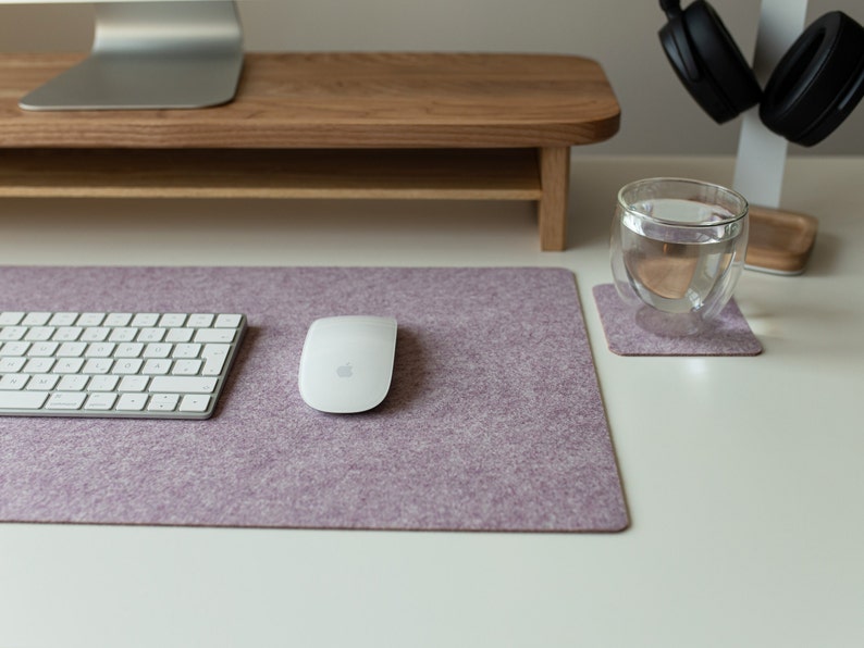 May include: A white wireless keyboard and mouse on a purple felt desk mat. The mat is on a white desk with a glass of water and a wooden coaster.