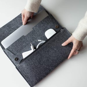 Könnte beinhalten: Eine graue Filz-Laptoptasche mit Reißverschlusstasche, die einen Laptop, ein Ladegerät, eine Maus und Kabel enthält. Die Tasche ist geöffnet und zeigt den Inhalt. Schlichtes und funktionales Design zum Schutz von Elektronik.