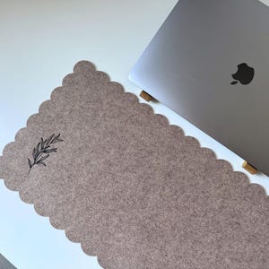 Puede incluir: Una alfombrilla de escritorio de fieltro color topo con borde festoneado y una ilustraci&oacute;n bot&aacute;nica negra. Un ordenador port&aacute;til plateado es parcialmente visible en el lado derecho de la alfombrilla, apoyado en soportes de madera. La alfombrilla est&aacute; sobre una superficie blanca.