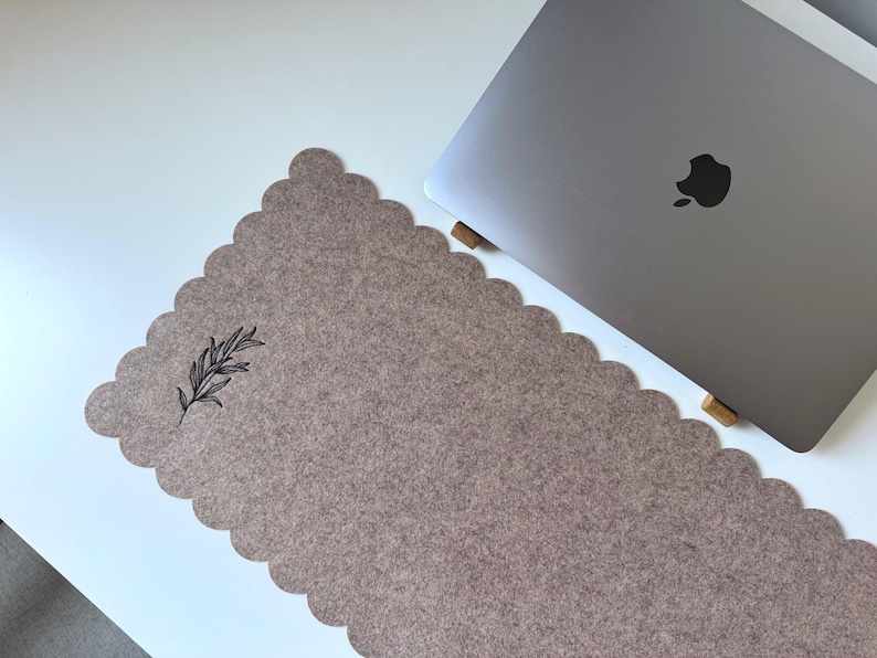 Puede incluir: Una alfombrilla de escritorio de fieltro color topo con borde festoneado y una ilustraci&oacute;n bot&aacute;nica negra. Un ordenador port&aacute;til plateado es parcialmente visible en el lado derecho de la alfombrilla, apoyado en soportes de madera. La alfombrilla est&aacute; sobre una superficie blanca.