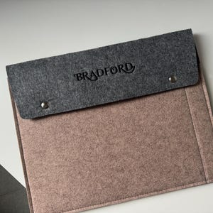 May include: A rectangular felt laptop sleeve featuring a dusty rose main body and a dark gray fold-over flap. The flap is secured with two silver-toned metal snaps and displays the word BRADFORD embossed in a dark, stylized serif font.