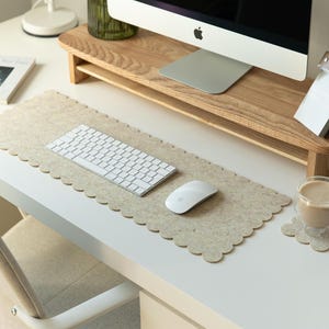 Könnte beinhalten: Ein moderner Schreibtisch mit einer weißen Tastatur, einer Maus und einer beigefarbenen Filz-Schreibtischunterlage mit gewelltem Rand. Ein Monitorständer aus Holz und eine Kaffeetasse sind ebenfalls zu sehen, was einen sauberen Arbeitsplatz schafft.